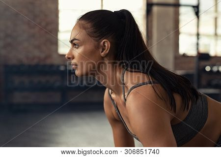 Young woman doing squat exercise while looking away at gym. Closeup of muscular girl while exercising in fitness center. Fit and concentrated woman doing strength workout.