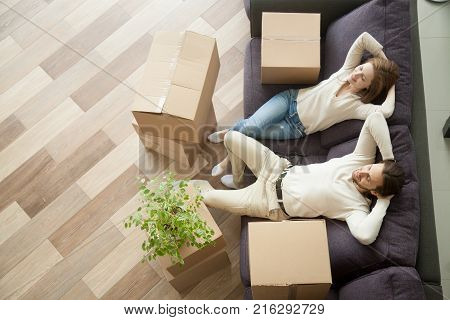 Couple resting on couch after moving in, man and woman relaxing on sofa just moved into apartment with cardboard boxes on floor, happy satisfied homeowners enjoying first day in new home, top view