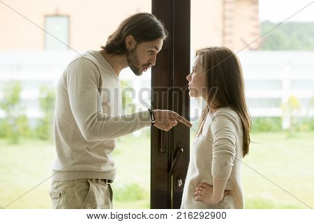 Unhappy young couple arguing standing at house door, angry husband pointing at wife blaming her of problems, conflicts in marriage, bad relationships, man and woman having quarrel or disagreement