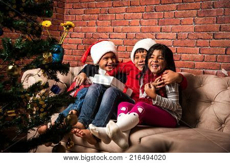 Indian kids celebrating christmas - cute little Indian kids playing, laughing and having fun with gift boxes on christmas, sitting on floor or sofa with christmas tree in the background