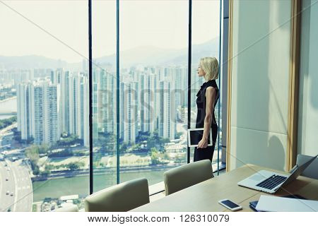 Blonde woman proud CEO is holding touch pad and looking in office window with cityscape while is standing in conference room near table with open laptop computer during her business trip in Hong Kong