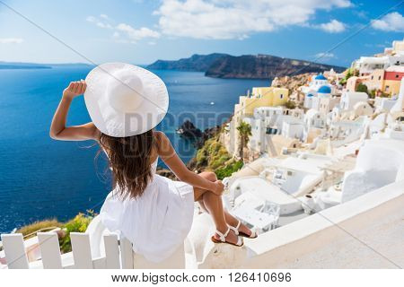 Tourist woman enjoying view of beautiful white village of Oia with Caldera and mediterranean sea. Young stylish female model wearing sunhat and red dress enjoying summer travel vacation in Europe.