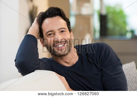 Portrait of a smiling happy young man resting while sitting on sofa. Handsome guy looking at camera. Young latin man in casual relaxing at home. Man relaxing on sofa with arm behind the head.
