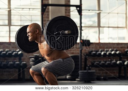 Determined african man lifting weight in gym. Muscular strong man taking efforts to lift weight barbell in fitness center. Black shirtless bodybuilder doing squatting with a barbell on shoulder.