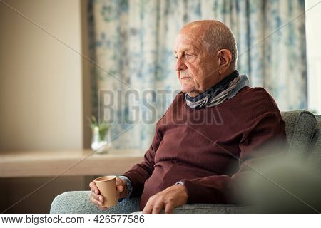 Depressed senior man sitting on armchair holding disposable cup of coffee and thinking. Frustrated retired man sitting on sofa. Sad mature man sitting alone at nursing home with sad expression.