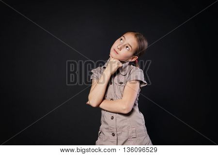 Thinking child portrait. Little preteen girl pensive, consider something, looking up. Primary school student solving some problem at black studio background with copy space.