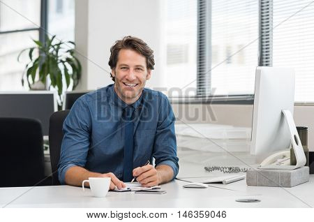 Cheerful young businessman working at desk in office. Successful business man sitting in office with a cup of coffee. Portrait of happy young businessman laughing and looking at camera.