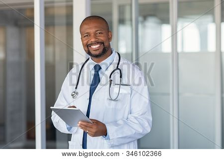 Portrait of confident mature black doctor consulting digital tablet and looking at camera. Smiling african american doctor with stethoscope using tablet at medical clinic. Happy healthcare worker.