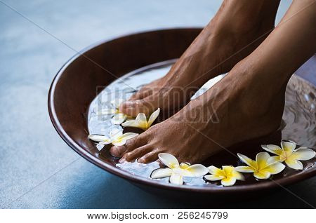 Woman soaking feet in bowl of water with floating frangipani flowers at spa. Closeup of a female feet at wellness center on pedicure procedure. Woman feet in spa wooden bowl with exotic white flowers.