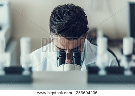 Young Scientist Using Microscope In Laboratory. Male Researcher Wearing White Coat Sitting At Desk A