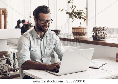 Young Smiling Man Working On Laptop In Cafe. Happy Handsome Bearded Business Man Sitting At Table An