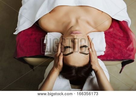 Attractive brunette woman getting massage spa treatment lying on a massage table, hands working on massaging woman's head. Relaxing facial massage at spa salon, top view.