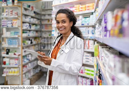 Happy friendly multiethnic pharmacist doing inventory in a provided and modern pharmacy while looking at camera. Portrait of smiling african doctor woman working in drugstore with digital tablet.