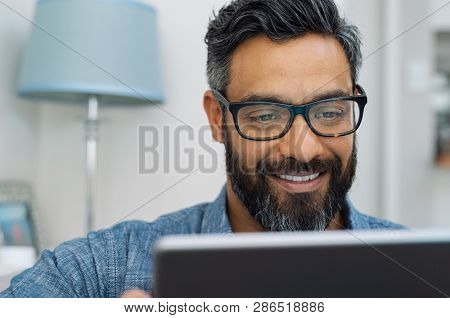 Relaxed mature man at home using digital tablet. Handsome hispanic man using laptop on sofa. Confident multiethnic guy with spectacles and beard using digital laptop.