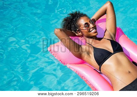 Beautiful african woman lying on pink mattress in outdoor swimming pool. Young black woman sunbathing in swimming pool with copy space. Happy girl in glamour bikini relaxing in pool with eyeglasses.