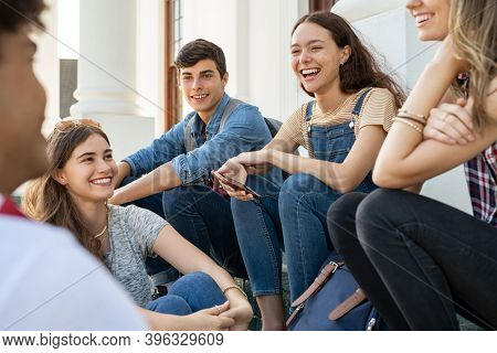 Group of happy young friends sitting in college campus and talking. Cheerful group of  smiling girls and guys feeling relaxed after university exam. Excited millenials laughing and having fun outdoor.