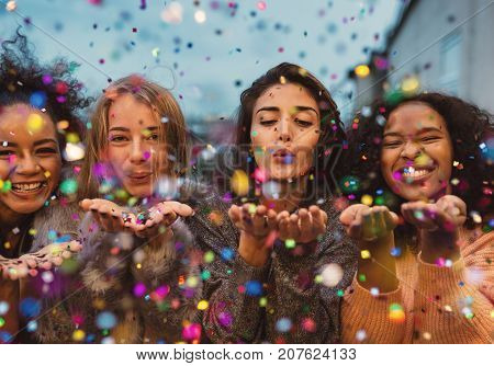 Young women blowing confetti from hands. Friends celebrating outdoors in evening at a terrace.