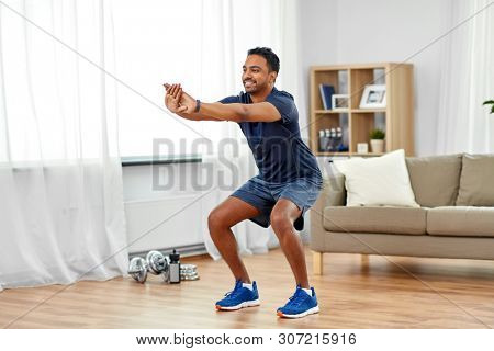 sport and healthy lifestyle concept - smiling indian man with fitness tracker exercising and doing squats at home