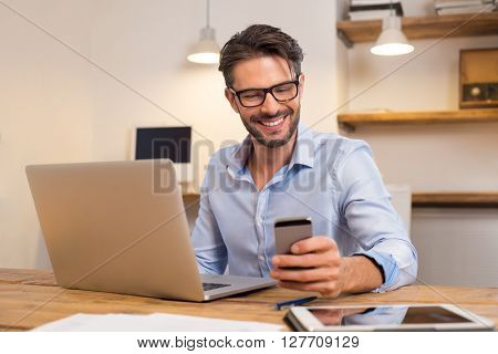 Young happy businessman smiling while reading his smartphone. Portrait of smiling business man reading message with smartphone in office. Man working at his desk at office.
