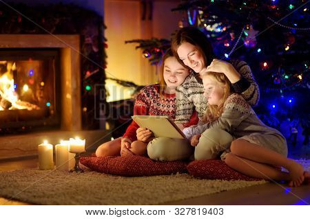 Mother And Her Two Cute Young Daughters Using A Tablet Pc At Home By A Fireplace In Warm And Cozy Li