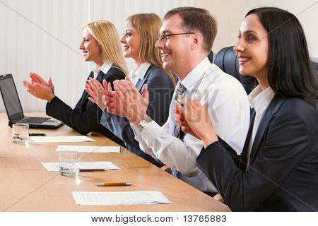 Portrait of successful applauding young people sitting in line in black comfortable chairs at the table with monitor, papers and glasses of water on it