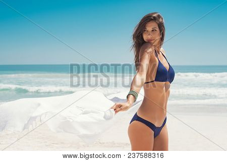 Young woman waving white scarf in wind at beach. Latin happy girl in blue bikini holding tissue and looking away at sea. Sexy fashion woman playing with the scarf on seashore. 