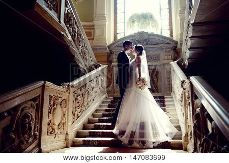 Wedding couple indoors is hugging each other. Beautiful model girl in white dress. Man in suit. Beauty bride with groom. Female and male portrait. Woman with lace veil. Cute lady and handsome guy