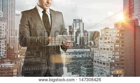 Double exposure image of a modern businessman with digital tablet over a city view. Business man with modern tablet with skyskrapers in background during the morning sunlight. New technology concept.