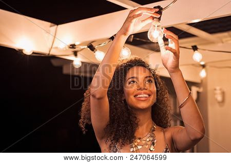 Cheerful young woman wearing party dress changing bulb light in patio. Happy smiling girl making preparation of party by adding lights outdoor. Happy beautiful woman enjoying fixing bulbs in backyard.