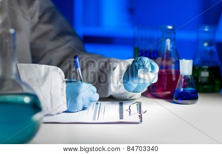 science, chemistry, medicine and people concept - close up of young scientist with chemical sample taking notes on clipboard and making test or research in laboratory