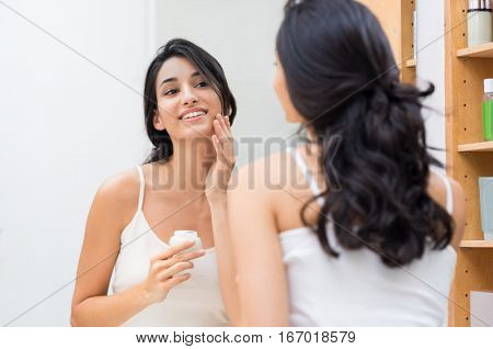 Woman caring of her beautiful skin on the face standing near mirror in the bathroom. Young woman applying moisturizer on her face. Smiling girl holding little jar of skin cream and applying lotion.