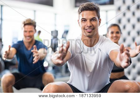 Group of happy people exercising in squat position. Man doing stretching exercise and looking at camera. Portrait of smiling personal trainer doing squat exercise with fitness class in background.