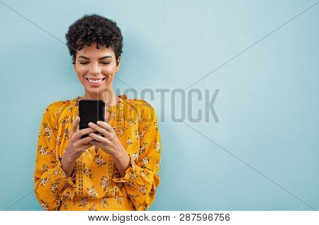 Happy smiling black woman using smart phone. Young brazilian woman writing a message with mobile isolated on blue background. Portrait of african american stylish girl using cellphone with copy space.