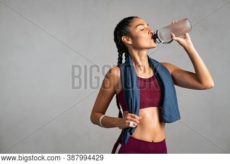 Fitness woman drinking water standing on gray background with copy space. Portrait of sweaty latin woman take a break after intense workout. Mid adult lady drink from water bottle after gym workout. 