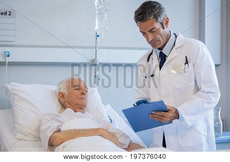 Mature doctor reading medical reports of a senior patient lying on bed at hospital. Doctor on a medical round while checking senior patient report. Professional doctor with clipboard at hospital ward.