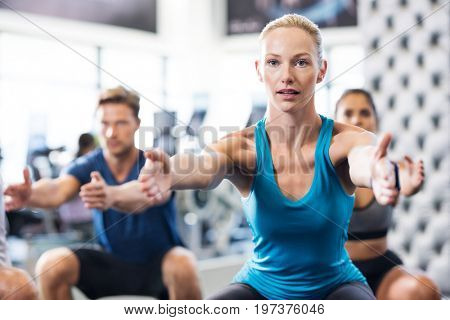 Young woman exercising in gym with people in background. Fit woman exercising with stretched hands and squats in modern gym. Fitness class exercising in gym.