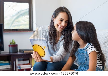 Happy mother reading story book to little girl. Happy latin mother and daughter reading a book while sitting on couch at home. Mother reading a fairy tale to adopted child.