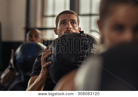 Portrait of young man lifting medicine ball with class in fitness center. Strong athlete doing strength training at gym. Guy holding heavy ball and doing squat exercise.