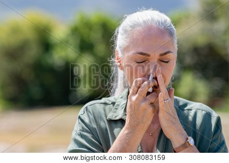 Senior woman having flu using nasal spray to help herself. Woman using a nasal spray at park for allergy. Ill mature woman with grey hair inhaling medicine against allergy.