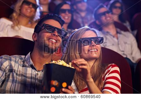 Happy couple sitting in movie theater, watching 3D movie, eating popcorn, smiling.