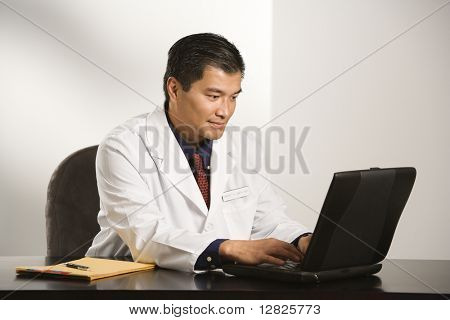 Asian American doctor hombre sentado en el escritorio con gráficos escribiendo en la computadora portátil.