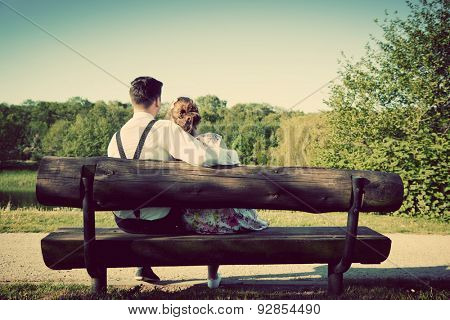 Young couple in love sitting together on a bench in summer park. Man wearing shirt with suspenders. Happy future, marriage concepts. Vintage