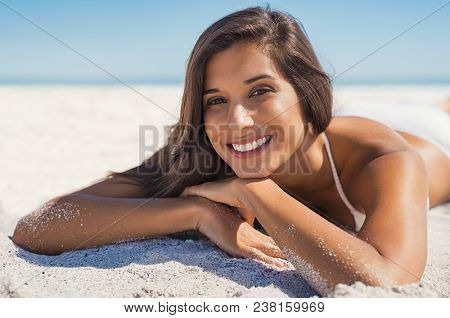 Happy young woman in swimwear lying on sand at beach. Portrait of smiling young woman relaxing on beach and looking at camera. Beautiful latin girl during summer vacation.