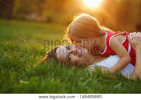 Mother and daughter lying on the lawn. Daughter kiss her mother on the cheek. Family in the city park outdoors. Happiness of motherhood and childhood.