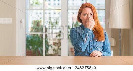 Redhead woman at home smelling something stinky and disgusting, intolerable smell, holding breath with fingers on nose. Bad smells concept.