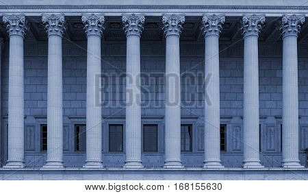 A building facade with corinthian columns in a row (colonnade). The neoclassical building style resembles a law court / courthouse, university, library or public administration building. Blue color tone.