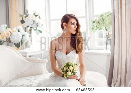 Wedding. Bride in beautiful dress sitting on sofa indoors in white studio interior like at home. Trendy wedding style shot. Young attractive caucasian brunette model like a bride against big window tender posing.