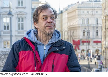 Street Portrait Of An Elderly Man 45-50 Years Old In A Jacket On A Blurry Background Of A European C