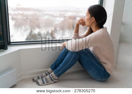 Depressed young girl feeling sad an lonely, anxious looking out the window in winter. Unhappy Asian woman alone, depression at home.