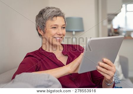 Smiling senior woman looking her digital tablet while sitting on sofa. Portrait of mature happy woman relaxing at home with digital tablet. Happy lady with gray hair browsing on laptop in living room.